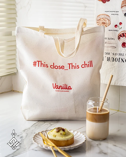 توت بگ اختصاصی سفید رنگ برای برند وانیلا، با متن : #This close_This chill و نام برند vanilla با رنگ قرمز روی توت بگ، روی میز کنار تت بگ یک لیوان قهوه و کیک هم قرار دارد